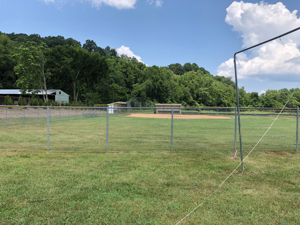 The baseball field at Creed Fields Park is shown from behind the outfield with a backdrop of mature trees on a sunny summer day.