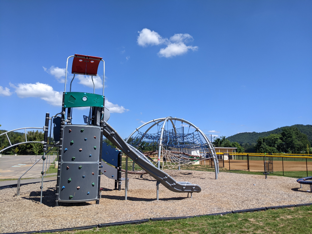 The playground at Creed Fields Park has a slide with a climbing wall and multiple climbing structures on a sunny summer day.