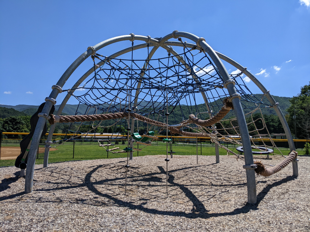 A giant modern climbing dome with many different types of rope is shown at the playground at Creed Fields Park on a sunny summer day.