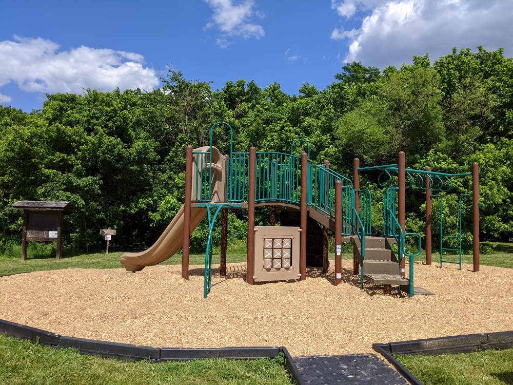 The playground at Eastern Montgomery Park has a mulch base and is shown in front of trees and the walking trail starting point.