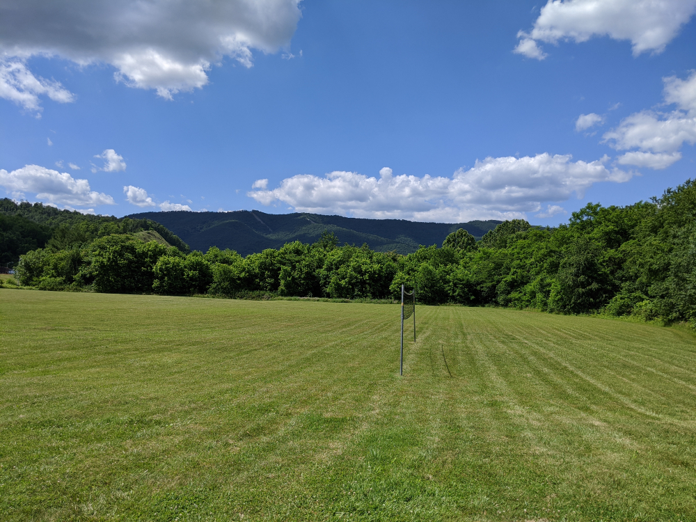 The open green space bordered by trees under the backdrop of mountains on a sunny summer day.