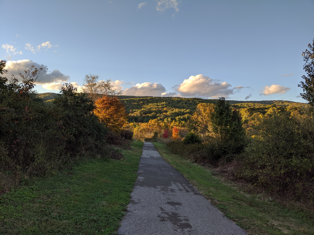 The Huckleberry Trail winds through Heritage Park towards the Gateway Trail in early fall, with a view of Brush Mountain in the distance under fluffy clouds illuminated with golden evening sun.