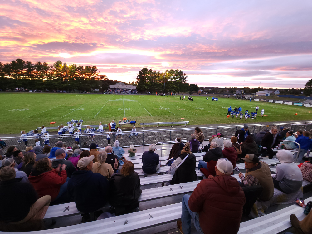 Fans watch a youth football game and cheerleaders to the backdrop of an orange and pink sunset.