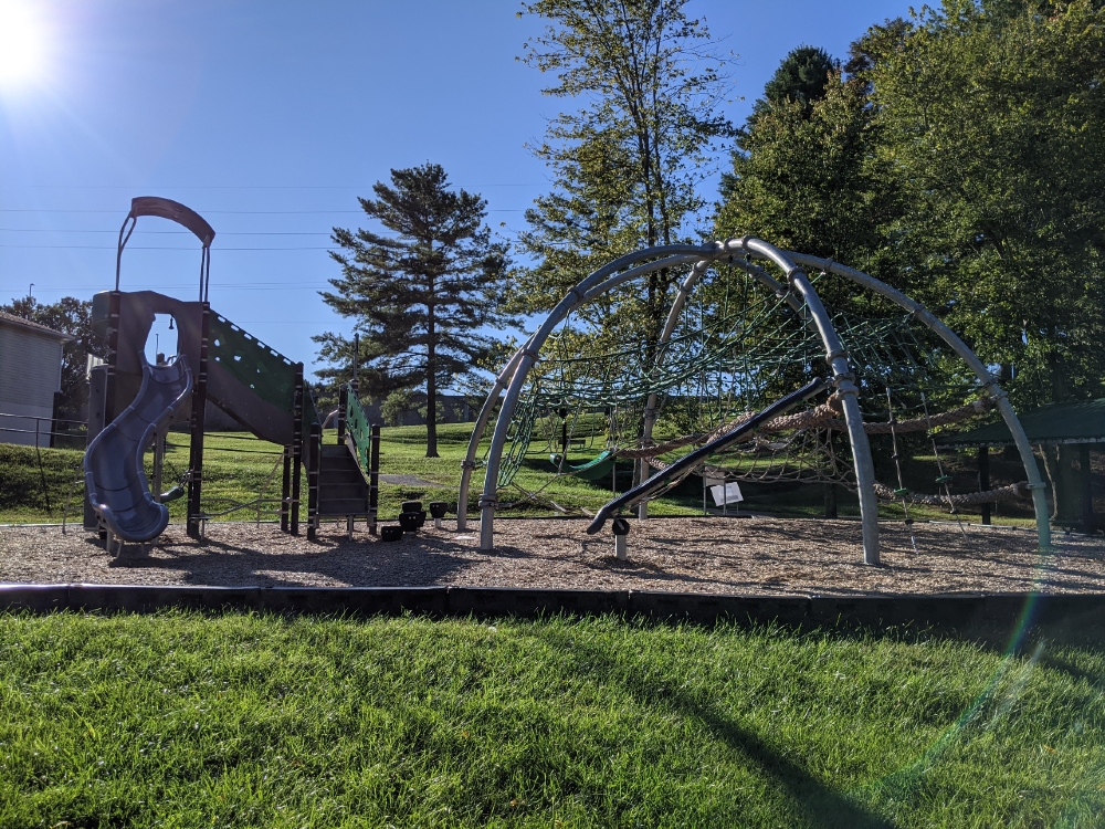 The playground at Mid County Park features a slide and large climbing areas, and has a mulch base surrounded by green space and trees.