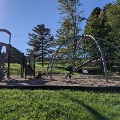 The playground at Mid County Park features a slide and large climbing areas and has a mulch base surrounded by green space and trees.